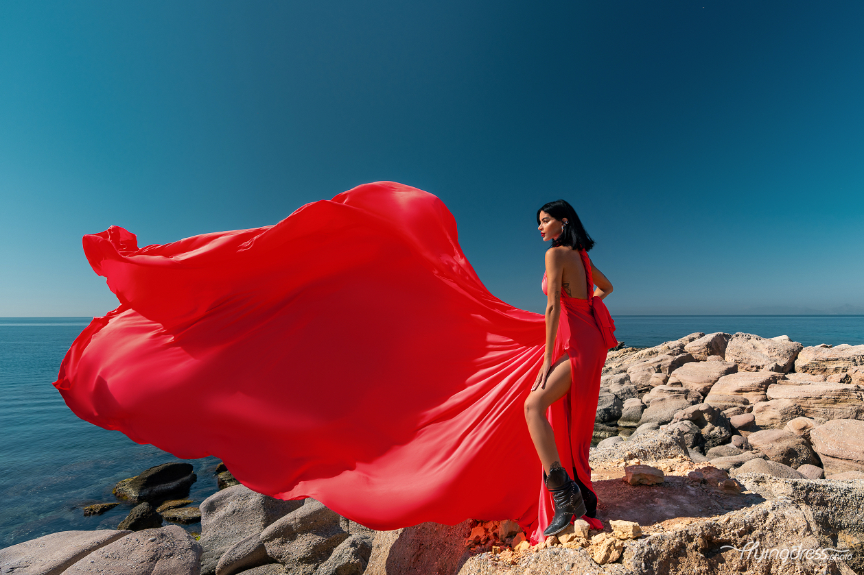 A woman in a flowing red dress and black boots stands confidently on rocky cliffs with the deep blue sea and clear sky of Kavouri, Athens in the background, creating a striking and vibrant scene.